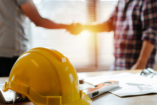 yellow hardhat with a deck builder and a happy customer shaking hands in the background over custom deck plans